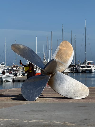 This media file features a large, metallic ship propeller prominently displayed in the foreground. The propeller has four blades, and one of them shows signs of wear and tarnishing. Behind the propeller, a person is visible, holding a smartphone in their outstretched hand as if taking a picture, and also holding a yellow object. The person appears to be standing on a dock or pier.

The background reveals a marina filled with sailboats and other vessels docked in calm water. The masts of the sailboats stretch upwards, some silhouetted against a clear, bright blue sky. The overall scene suggests a sunny day. The reflection of the boats in the water is visible, adding depth to the image. The texture of the ground in the foreground suggests it is a paved surface, possibly concrete or asphalt, with some wear and tear.

The orientation of the image is rotated, with the sky and water appearing on the right side and the ground and propeller on the left. The lighting indicates it is likely daytime, with the sun casting shadows. There is no visible text in the image. The setting is a marina, and the activity appears to be a person taking a photograph of themselves or the surroundings with a ship propeller as a prominent foreground element.