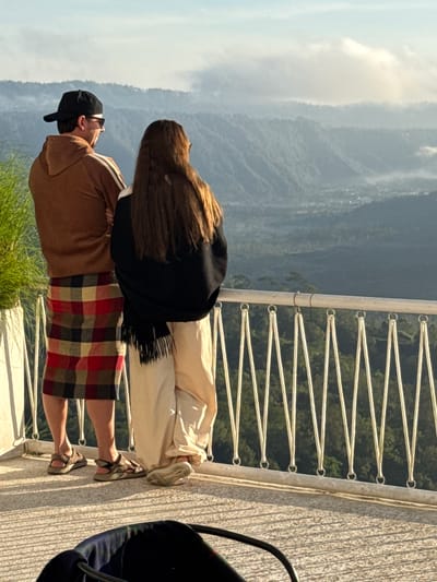 Two individuals stand on a balcony overlooking a mountainous landscape in Kedisan, Indonesia. One person, wearing a brown jacket and a red and black plaid skirt, looks out at the scenery. The other person, standing closer to the railing, has long brown hair, wears light-colored pants and a black shawl. The balcony features a white metal railing with a zigzag wire design. Greenery is visible at the base of the balcony. In the foreground to the right is a dark object partially visible. The backdrop showcases layers of mountains fading into the distance. The sky is overcast.