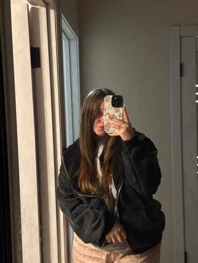 A female individual is depicted in a mirror selfie taken indoors in the city of Tlustenkhabl, Russia. The subject, positioned centrally, has long, straight, dark brown hair with lighter highlights, parted roughly in the middle. She is wearing a grey crew-neck t-shirt underneath an unzipped, dark grey or black full-zip hoodie with a faded appearance. Light pink or peach-colored patterned pants are visible on her lower body. Her right hand holds a smartphone, obscuring the lower half of her face. The smartphone has a beige or cream-colored case with a brown botanical pattern and appears to be an iPhone model with three visible rear camera lenses. A gold-colored ring is present on her left ring finger. Her left eye is visible, looking towards the reflected camera. The background, also reflected in the mirror, consists of light-colored walls and two light-colored door frames with vertical paneling, suggesting an interior room. The lighting is diffused, illuminating the subject and the surrounding area.