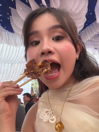A woman with dark hair, fair skin, and full pink lips is holding a wooden skewer with what appears to be grilled meat on it. She has an open mouth and is looking directly at the camera. She is wearing a light-colored, possibly tulle, garment and a gold necklace with rings as pendants. In the background, other individuals are present, but they are out of focus. There is a white and blue decorative element in the background, along with a white vertical ribbed or pleated structure. The location is Tay Son Commune, Vietnam.