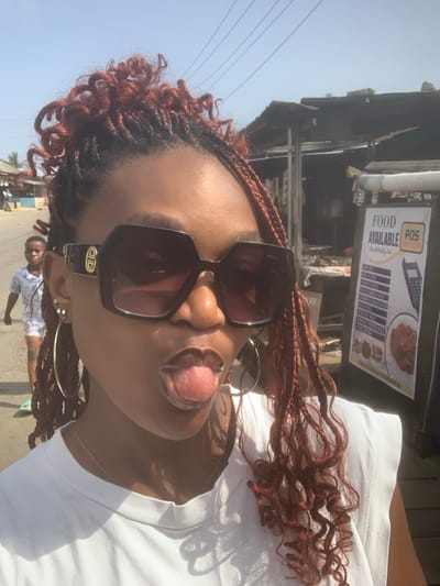 A close-up image shows an adult female in Anantigha, Nigeria, posing for a selfie. Her complexion is dark, and her hair is styled in reddish-brown braids, partially gathered upwards and loose over her shoulders. She wears large, dark-tinted rectangular sunglasses with a visible Gucci logo on the temple, silver-toned hoop earrings, multiple ear studs, and a thin silver chain necklace over a white sleeveless garment. Her tongue is protruding from her mouth.
In the mid-ground, a young dark-skinned male child walks away from the camera on what appears to be an unpaved road. To the right, a weathered market stall with a corrugated metal roof stands beside a white signboard advertising "FOOD AVAILABLE" and "POS SERVICES." The signboard lists food options including "Rice & Stew," "Beans & Plantain," "Yam Porridge," "Eba & Soup," "Amala," "Pounded Yam," "Moimoi," "Akara," "Egusi," "Okra Soup," and "Chicken Pepper Soup," and features an illustration of a POS terminal. Overhead, power lines span across a clear sky. Further down the street on the left, additional structures with various roofing are visible.
