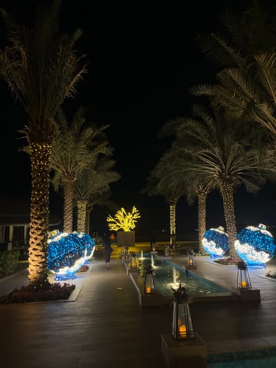 The image depicts an illuminated outdoor scene at night within Ras Al Khaimah, United Arab Emirates, characteristic of a luxury resort or similar establishment. The foreground and midground feature a paved pathway (dark wood or large tiles) flanked by numerous tall palm trees, their trunks wrapped with warm white string lights.
On the right side of the pathway, three large, spherical, ornamental structures are visible, composed of blue and white lights in swirling patterns, resembling oversized festive baubles. A similar blue and white ornamental sphere is partially visible on the far left. A narrow, rectangular water feature with multiple small fountains runs parallel to the pathway, containing light blue water. Several dark-colored decorative lanterns, containing glowing light sources (simulating candles), are positioned along the edges of the water feature and the pathway.
In the midground, a solitary individual is walking away from the viewer on the pathway. The person is dressed in a dark, long garment and carries a small, light-colored handbag (possibly orange or tan) over their left shoulder. Beyond this individual, a prominent, abstract, tree-like or coral-like sculpture, brightly illuminated in yellow/gold lights, stands on a raised platform.
Further in the background, to the far left, a portion of a building with warm interior lighting is visible, suggesting a structure such as a restaurant or reception area. The sky above is dark and clear, indicating nighttime conditions. Distant, indistinct lights are visible beyond the main decorative elements, suggesting further development or possibly a body of water.