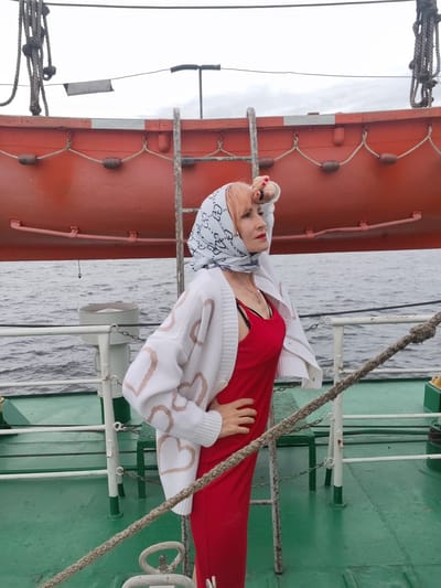 A woman with pink hair stands on the deck of a ship, looking out to sea. She is wearing a red dress and a white cardigan with a heart pattern. A white scarf with a black pattern is tied around her head. The ship has an orange lifeboat mounted above her. The sky is overcast, suggesting a cloudy day. The water is choppy, indicating the ship is in motion. The deck is painted green, with white railings. A thick rope is strung along the railing. The woman has her hand on her hip and appears thoughtful. The scene is likely taking place in Bereg, Russia, given the context. There is no visible text in the image.