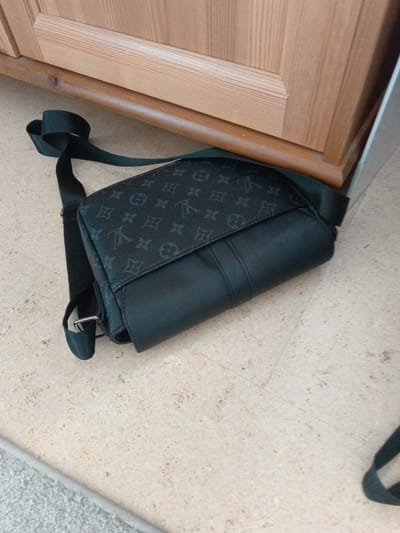 A black sling bag with a monogrammed pattern lies on a light-colored tiled floor. The bag is positioned diagonally, with its strap extended. The monogram is subtle, appearing as darker shades of black on the fabric. The bag has a flap closure and a solid black section on the lower front. A light wooden cabinet or furniture is visible behind the bag, taking up the top portion of the image. The lighting suggests an indoor setting, possibly during the daytime. No people are visible in the frame. The overall scene is still and focuses on the object. The visible text is limited to the repeating monogram on the bag, which appears to be the Louis Vuitton pattern. The image is taken from a high angle, looking down at the bag.