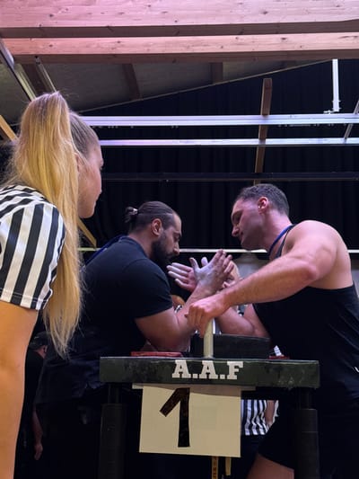 This image depicts an indoor arm wrestling competition taking place in Reisenberg, Austria. Three individuals are prominently featured. On the left, a blonde female referee with her hair in a high ponytail is partially visible from the chest up, wearing a black and white striped shirt and observing the competitors. In the center, two muscular adult males are positioned at a dark green arm wrestling table, actively engaged or preparing to engage in a match. The male on the left has a dark beard and hair tied in a bun, wearing a dark short-sleeved shirt. His hands are clasped with his opponent's, both coated with white chalk. The male on the right has short dark hair and is wearing a dark tank top with a blue strap visible around his neck; his hands are similarly chalked and clasped with his opponent's. A smaller, indistinct figure is partially visible behind their clasped hands. The arm wrestling table displays white lettering "A.A.F" on its front panel, below which a white sign with a large black number "1" is affixed. Red pads are visible on the table surface under each competitor's elbow area. The background consists of dark drapes or walls. Overhead, exposed wooden beams and metal support structures are visible, indicative of a building's ceiling.