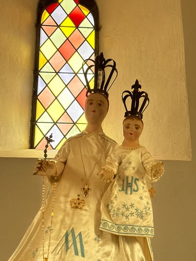 Two statues, likely of the Virgin Mary and the Christ Child, are positioned in front of a stained-glass window in Bech, Luxembourg. The statue on the left, representing the Virgin Mary, is larger and wears a long, satin robe adorned with a cross pendant and a crown. She holds a rosary in her left hand. The statue on the right, presumed to be the Christ Child, is smaller and also wears a satin robe with embroidered stars and the monogram "JHS". It is also crowned and holds a small cross. The stained-glass window behind them features a diamond pattern of red, yellow, and white panes, illuminated by light. The surrounding wall is a light-colored, textured surface.