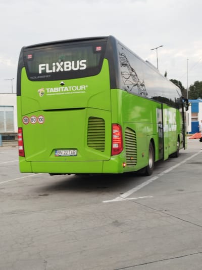 This is a medium shot, exterior, low-angle photograph of the rear of a bright green FlixBus tour bus. The bus is parked in what appears to be a parking lot with cracked concrete and white lane markings. The bus has "FLIXBUS" written in large white letters on the back windshield and "TABITATOUR" with a logo above it. There are speed limit signs indicating 50, 80, and 100. The license plate reads "BN 22 TAB". In the background, there is a building with blue and white accents, and some trees. The sky is overcast. There are no people clearly visible in the immediate vicinity of the bus. The location is specified as Tânganu, Romania.
