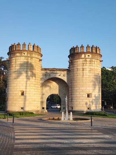 The image shows the Puerta de Palmas, a historic city gate in Badajoz, Spain. The gate is made of stone and features two imposing circular towers with crenellated tops, connected by an arched passage. A fountain with water spraying upwards is located in the foreground, in front of the gate. The ground is paved with interlocking stones, and a pedestrian crossing is visible, with alternating dark and light gray stripes.
The scene is illuminated by the warm, golden light of the setting sun, casting long shadows and highlighting the texture of the stone. The sky is clear and a vibrant blue, indicating fair weather. Trees with lush green foliage frame the sides of the gate, and a white car is seen passing through the archway in the background. Two people are walking on the right side of the image, suggesting a normal day in the city.
The overall mood is peaceful and serene, capturing the grandeur of the historical monument bathed in the soft light of late afternoon or early evening. The architectural details of the Puerta de Palmas, including the small windows and decorative rope molding around the towers, are clearly visible. The image conveys a sense of timelessness, blending the historical significance of the gate with the everyday life of the city. No visible text is present in the image.