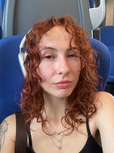 This close-up selfie captures a young woman with prominent reddish-brown, curly hair, looking directly at the camera. Her expression is calm and relaxed, with a neutral mouth and soft gaze. She has a natural makeup look, featuring a subtle blush and pink-toned lips. She is wearing a black strap top or bra, and layered delicate gold necklaces with small pendants are visible on her chest. A light-colored (possibly silver or white) headphone band or similar accessory is visible near her left ear, partially covered by her hair.
The setting appears to be the interior of a vehicle, likely a train or bus, indicated by the blue upholstered seat or headrest she is resting against. Behind her, parts of grey and white plastic-like structures, possibly armrests or seat backs of adjacent seating, are visible. The bright lighting suggests it is daytime. The image conveys a sense of travel or relaxation. No text is visible within the image. The image is set within Stara Pazova Municipality, Serbia.