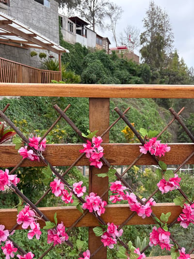 This image depicts a close-up view of a wooden fence adorned with artificial pink flowers and green leaves. The fence is constructed with horizontal wooden planks and a diamond-shaped lattice made of thinner, darker wooden sticks. Behind the fence, a lush green hillside is visible, dotted with various types of vegetation and some scattered yellow flowers. In the background, buildings are situated on the slope, with one structure featuring a gray, textured facade and a wooden balcony. Another building with a red roof and a flag is also present on the upper part of the hill. The sky is overcast and gray. The scene appears to be an outdoor setting, likely a residential area with a garden or terrace. The image was taken in San Vicente de Azpitia, Peru.