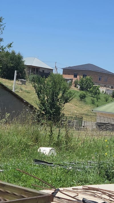 A slightly overcast daytime scene in Leñgir, Kazakhstan, depicts a rural residential area. In the foreground, tall green grass and weeds are interspersed with a wire fence. A white plastic container is partially visible amongst the vegetation. Beyond this, a gently sloping grassy hill is dotted with several trees and at least two houses. One house, on the left, has a light-colored, angular roof and a darker base. Another, larger house, is situated further up the hill on the right, constructed from brick with a dark, pitched roof. A few smaller buildings and structures are also present on the hillside. The sky is a clear, light blue.