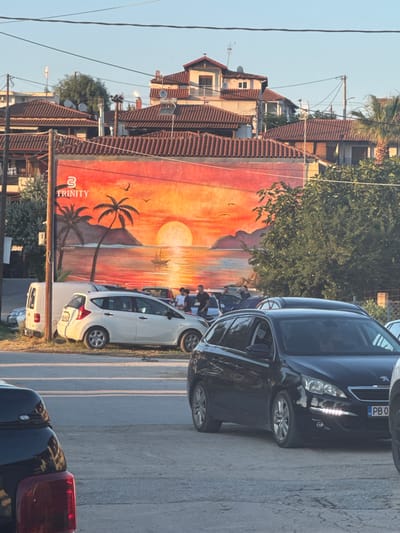 The image depicts a street scene in Ammouliani, Greece. Dominating the background is a building adorned with a large mural of a sunset over the sea, featuring palm trees, islands, and a sailboat. The mural has the word "TRINITY" prominently displayed with a logo above it. In the foreground, several cars are parked along the roadside. A white hatchback is parked to the left, next to a white van. A black station wagon is parked in the center, facing right, with its headlights illuminated. Another black vehicle is partially visible in the lower left corner. A few individuals are standing near the white hatchback, some appearing to be interacting. The environment suggests an outdoor setting, likely during the daytime with clear skies, as indicated by the bright sunlight and shadows. The presence of parked cars, buildings, and overhead wires points to a developed urban or semi-urban area.