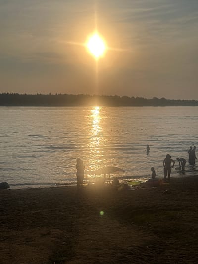 This image depicts a serene sunset scene at the beach in Ust-Kachka, Russia. Silhouetted figures are visible wading in the calm, shimmering water, reflecting the warm, golden hues of the setting sun. The sky is a soft gradient of orange and grey, with the sun positioned high, casting a bright, starburst-like glow. A line of buoys stretches across the water in the distance. The foreground is a sandy beach, also in silhouette, with a few scattered beach toys and an umbrella. The overall atmosphere is one of peaceful relaxation and enjoyment of the natural beauty of the location.