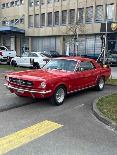 The image shows a red 1965-1966 Ford Mustang parked along the side of a street, likely in Wetzikon (ZH), Switzerland, given the license plate "ZH". The car is parked next to a building with the name "KET TIGER" and a logo featuring a lion. There is a white Audi SUV and a white Volkswagen pickup truck parked further down the street, adorned with small flags bearing the same lion logo. A yellow painted stripe runs parallel with the curb. The building's architecture suggests an industrial or commercial area. The scene appears to be in daylight. There are no visible people.