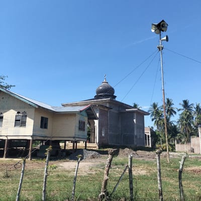 A daylight exterior shot captures a tranquil rural scene in Tanjongkeumala, Indonesia, featuring a mosque, a traditional stilt house, and a prominent loudspeaker tower against a clear blue sky.
In the center-right of the frame, a mosque with concrete and brick walls is visible, topped by a distinctive black and gold patterned dome. The dome is crowned with a crescent moon and star finial. Piles of gravel near the mosque's base suggest ongoing construction or renovation. To the left of the mosque stands a weathered, light-colored wooden stilt house featuring louvered windows and a corrugated metal roof, supported by elevated wooden pillars.
A tall, slender metal pole, likely serving as a minaret substitute or a dedicated broadcast tower for the Adhan (call to prayer), rises on the right side. It is equipped with several horn-shaped loudspeakers mounted at the top, along with metal climbing rungs and stabilizing guy wires. Behind the mosque and pole, numerous palm trees are scattered across the landscape, indicating a tropical environment.
The foreground is marked by a rustic fence constructed from rough wooden posts and wire. The ground consists of a mix of dry grass, bare earth, and small gravel piles. The photograph was taken on a bright, clear day, with strong sunlight casting noticeable shadows, suggesting a time between late morning and mid-afternoon. No people or visible text are present in the image. The scene portrays a quiet community setting centered around the mosque.