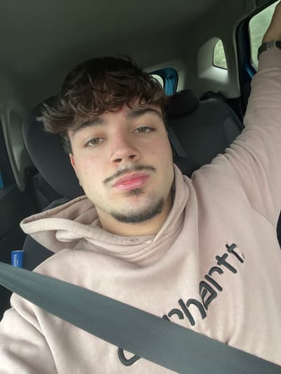 A young adult male with light skin, brown eyes, dark brown curly hair, a mustache, and a chin strap beard is seated inside a vehicle, located in Lamego, Portugal. He wears a light peach-colored hooded sweatshirt with partial black "Carhartt" branding and a dark grey seatbelt diagonally across his chest. A silver wristwatch is visible on his left wrist, and his right arm is raised, with the hand out of frame. The vehicle's interior features dark seats and trim, with a blue-framed rear side window revealing blurred greenery outside. A blue object, possibly branded "Sparco Corsa," is visible near the left side of the seat. The individual looks directly at the camera.