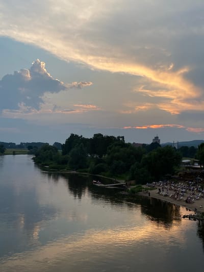 This image captures a sunset scene over the Weser River in Rinteln, Germany. The sky is filled with dramatic clouds, with a striking, luminous cloud formation on the left side and streaks of orange and yellow light illuminating the sky on the right. The river, wide and calm, reflects the colors of the sky. On the right bank of the river, a sandy beach area is populated with numerous people relaxing on lounge chairs under umbrellas. A beach bar or cafe structure is visible behind the seating area. Adjacent to the beach, a dense line of trees forms a natural backdrop. In the distance, a tall, white tower-like building is visible above the trees, along with other structures and hills. Two small boats are in the water near the shore on the left side of the frame. The overall atmosphere is one of leisure and natural beauty during a sunset.