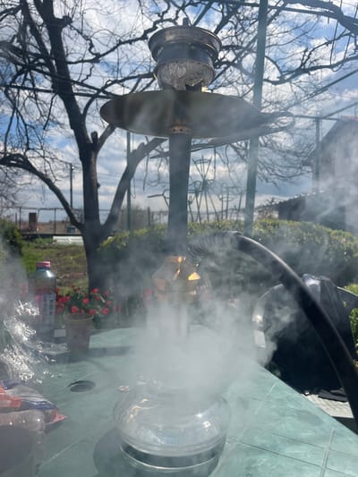 The image shows a hookah on a green plastic table outdoors in what appears to be Hrishteni, Bulgaria. The hookah is actively emitting smoke. In the background, there are bare tree branches and a garden with red flowers. Some discarded packaging lies on the table around the base of the hookah. The environment suggests a residential outdoor space.
