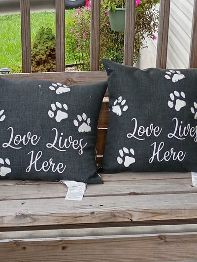 Two dark gray throw pillows with white paw prints and the words "Love Lives Here" are displayed on a weathered wooden bench. The pillows are patterned with multiple white paw prints. The text is in a cursive font. Behind the bench, a wooden railing separates the viewer from a grassy area with a small evergreen tree and pink flowers. The scene appears to be outdoors, possibly on a porch or deck. The location is specified as Lawrenceburg, United States of America, based on the file name.