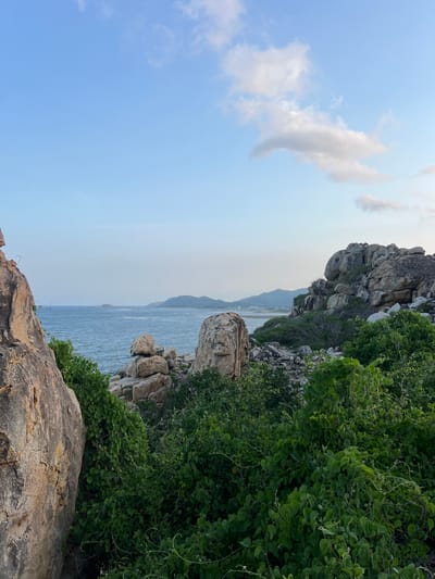 The image depicts a coastal landscape in Vĩnh Hải, Vietnam. The sky in the upper portion is light blue with several scattered white cumulus clouds, primarily situated on the right side. A defined horizon line separates the sky from a body of calm blue water, which occupies the central midground. In the distance across the sea, a small, low-lying island is visible towards the left. Further along the far horizon, a long, low mountain range is present, with faint outlines of structures discernible along its base. The foreground and midground are heavily textured with numerous large, irregularly shaped boulders and rock formations, displaying varying shades of brown and grey. Dense green foliage, consisting of shrubs and vines, covers the ground and intertwines amongst these rock formations. No human figures or animals are visible within the scene. The natural daylight suggests either morning or late afternoon conditions.