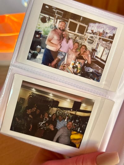 The image displays a close-up of an open white plastic photo album held by a hand with pink manicured nails. The album contains two instant photographs, indicative of Instax or Polaroid film, presented in clear plastic sleeves.
The top photograph, from 3-Y Uchastok, Russia, depicts four individuals posing in what appears to be an outdoor or semi-outdoor kitchen setting. On the far left, a shirtless, muscular man with a beard and short hair, wearing blue shorts or a towel, smiles and gives a thumbs-up with his left hand while embracing a woman to his right. This woman has long blonde hair, wears a light pink short-sleeved top and denim shorts, and has a tattoo visible on her left thigh. To her right, a shirtless man with short hair smiles at the camera. On the far right, a woman with dark hair wears a dark bikini top and makes a peace sign. In the foreground, a table covered with a patterned cloth holds multiple bottles (including beer), a juice carton, and other items. The background reveals a rustic kitchen with white wall cabinets, open shelving filled with various bottles and objects, a round mirror, and a large green watermelon on the floor to the left. The scene suggests a casual gathering.
The bottom photograph, also from 3-Y Uchastok, Russia, shows a group of at least eight adults in what appears to be an indoor bar or restaurant setting. The individuals are clustered together, smiling and laughing. On the far left, a woman in a dark patterned dress smiles. Several women in the center also smile, one wearing a black and white patterned top. To the right, an older man with grey hair in a light blue checkered shirt smiles, standing behind a seated man in a checkered shirt who laughs with an open mouth. The background features a well-stocked bar with numerous bottles on shelves, illuminated from behind. Decorations, possibly streamers, are visible hanging near the ceiling, suggesting a celebratory event.
The immediate surroundings outside the album are out of focus, with an orange-yellow object visible in the blurry background to the left.