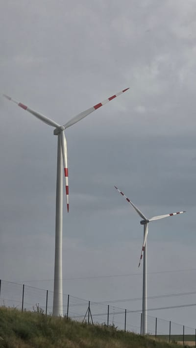 The image shows a view of two wind turbines in Poysdorf, Austria. The sky is overcast and cloudy. The wind turbines are tall, white structures with three blades each. The blades of both turbines have red and white stripes. The foreground consists of a grassy hill with a wire fence along the top. Power lines are visible in the background.