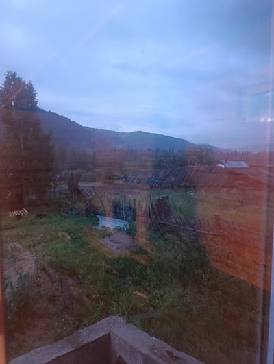 The image depicts a rural landscape at dusk, likely in the village of Birikchul, Russia. In the foreground, a concrete structure is visible, possibly part of a foundation or a well. Beyond this, an overgrown area with tall grass and some scattered dirt mounds leads to a dilapidated wooden fence and an assortment of wooden structures, likely sheds or outbuildings with damaged roofs. In the midground, more wooden structures with corrugated metal roofs are present, some appearing to be houses with chimneys. A dense line of trees forms the background, with a misty mountain range visible beyond. The sky is overcast with a pale blue hue, suggesting twilight or early dawn. The overall impression is one of rural neglect and the quiet stillness of evening.