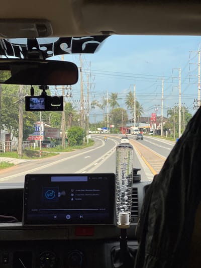 The image shows the interior of a vehicle, likely a van, looking out through the windshield. The dashboard features a large touchscreen display showing a music player interface. To the left of the display, a dashcam is mounted, with its screen showing a view of the road ahead. The interior also includes a gear shift knob filled with a clear liquid and bubbles. Outside the vehicle, a road curves to the right, with traffic signs and power lines visible. Palm trees and lush vegetation line the roadside, characteristic of a tropical environment. The sky is clear and blue. The scene appears to be captured in Tambon Sai Thai, Thailand, as suggested by the presence of Thai text on a sign. The overall impression is a snapshot of a journey on a sunny day in a Thai locality.