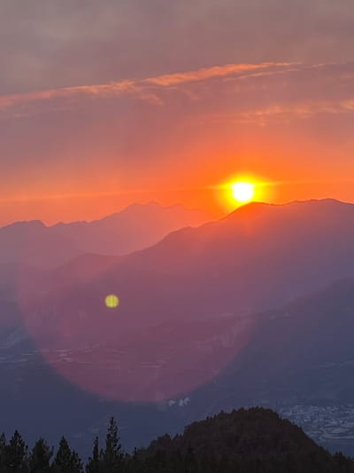 This image captures a sunset over a mountainous landscape, likely in or near San Juan de Plan/San Chuan de Plan, Spain, given the context. The sun, a bright, glowing orb, is partially obscured by the silhouette of a mountain range in the mid-ground. The sky is a vibrant gradient of orange and pink, with thin, wispy clouds streaking across the upper portion. The mountains themselves are layered, with the furthest peaks appearing as faint, hazy outlines due to atmospheric perspective. A distinct lens flare, appearing as a yellow circular halo, is present in the lower mid-ground. In the foreground, the dark silhouettes of trees and a forested hillside are visible, with a faint glimpse of a village or town nestled in the valley below on the right side of the image. The overall atmosphere is serene and dramatic, typical of a mountain sunset.