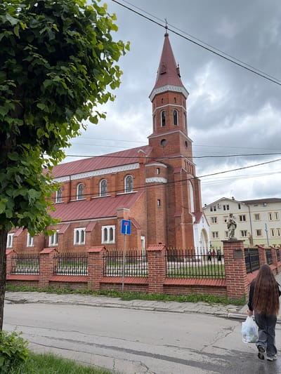 This is a full, eye-level shot of a large, brick church with a red-tiled roof and a tall, red-spired bell tower. The church is located on the left side of the frame, and the street is in the foreground. To the right of the church is a light-colored, multi-story building with many windows. A person with long, brown hair is walking away from the camera on the sidewalk, carrying a white plastic bag in their right hand. There is a black metal fence with gold accents in front of the church. The sky is overcast.