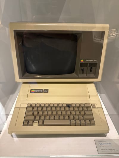 This media file shows an Apple Computer IIe, a classic personal computer from the early 1980s, displayed in a museum setting. The computer is cream-colored, with a separate monitor and keyboard unit. The monitor is on the left, displaying a dark screen, and has controls labeled "monitor ///", "CONTRAST", and "BRIGHTNESS" with accompanying knobs and a switch. The Apple logo, a rainbow-colored apple, is visible on the monitor's bezel. The keyboard unit is on the right, featuring a full QWERTY layout with additional function keys like "Reset", "Delete", and "Enhanced". The "apple IIc" logo is prominently displayed on the keyboard unit, also with the iconic rainbow apple.
A small informational plaque is attached to the right side of the display. The text on the plaque reads: "Computerrevolution", "Apple Computer lle", "mit Floppy-Diskettenlaufwerk", "Apple Inc. Corp., Cupertino, 1982", and "Historisches Museum Bern; -Inv. 61528". This indicates the computer is part of an exhibition on the "Computerrevolution" at the Historical Museum in Bern, Switzerland, and it includes a floppy disk drive. The lighting suggests an indoor, possibly artificial light environment, common for museum displays. The overall impression is one of historical preservation and exhibition of vintage technology.