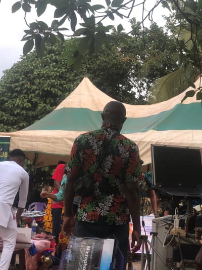 The scene is set outdoors in Awka Etiti, Nigeria. An adult male is prominently featured in the foreground, viewed from the back. He is bald and wears a black, short-sleeved collared shirt with a repeating pattern of green and orange/red leaves. A black pouch is slung across his back. He stands in front of a large canopy tent, which is cream or off-white with a green stripe. The tent's pointed peak is visible in the upper mid-ground.

To the left of the main subject, a person dressed in a white long-sleeved garment stands near a table laden with items. These items include several bottles, a small charcoal grill, and a tray holding what appear to be food items, possibly plantains. A white box is also on this table. Further left, dense green foliage is visible.

To the right of the main subject, an open black HP laptop is positioned on a stand or table. Adjacent to the laptop, a white power strip or multi-socket charger is connected to several cables. Below the laptop setup, a cardboard box labeled "Professional Wireless Microphone" is partially visible. Additional individuals are partially visible in the mid-ground behind the tables and equipment, some wearing patterned clothing or a red top. The background consists of dense green trees and foliage under an overcast sky.