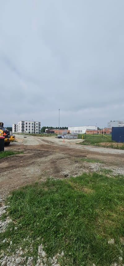 The image shows a construction site on a cloudy day in Maardu, Estonia. In the background, there are several modern apartment buildings with white facades and balconies. To the right of the apartment buildings, there is a long, low-rise industrial or commercial building with a white facade and some graffiti. Further to the right, there is another multi-story building with a grey facade. In the foreground, there is a dirt and gravel road with tire tracks, indicating recent vehicle activity. A yellow construction vehicle, possibly a roller, is partially visible on the left side of the frame, next to a green trash bin. Pallets of construction materials, likely pavers or bricks, are stacked on the gravel. Yellow caution tape is strung along the perimeter of the construction area. The overall scene depicts an active construction zone with ongoing development of residential and commercial properties.