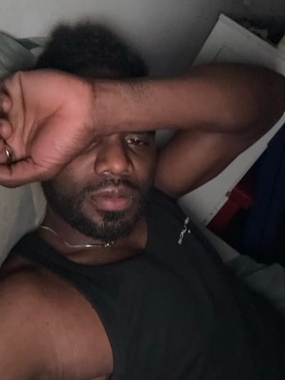 The image shows a man lying down, with his right arm covering his face, in what appears to be a bedroom in Aulnay-sous-Bois, France, located at 48.924 latitude and 2.499 longitude. The man is wearing a black tank top with a small white logo visible on the right shoulder strap. The image is taken from a low angle, making it difficult to see the full extent of the room. There is a white wall and a green blanket visible in the background. The man's face is obscured by his arm, but he appears to have dark skin, short black hair, and a short beard. His left arm is lying on a white sheet that is likely part of a bed. There is a light grey blanket behind his head. The room appears dimly lit, with light coming from an unknown source. There are no other people or objects visible in the image.  The man is not interacting with anything in the image.  He is lying down, seemingly asleep.