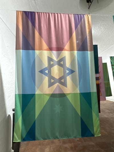 A rectangular flag displays a six-pointed Star of David at its center, overlaid on a muted, gradient-filled geometric pattern. The left side transitions through shades of purple and pink, while the right side features various tones of green. A circular haze surrounds the star. A smaller, star-like shape is visible in the green section to the right of the Star of David. The flag is hanging in an indoor setting with white and brick walls.