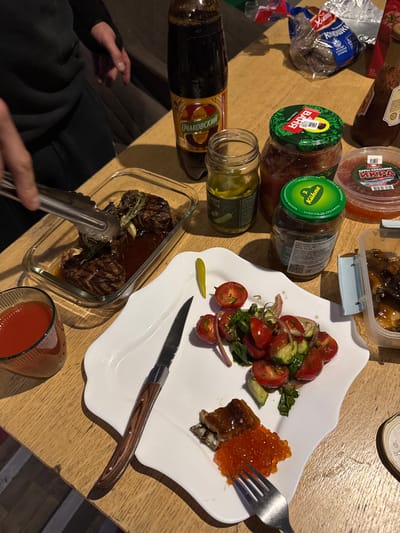 The image shows a table setting with various food items and drinks. On the table, there are jars of pickles, a container of what appears to be red caviar, a container of stewed food, and a loaf of bread in a plastic bag. A white plate contains a salad of tomatoes, cucumbers, and red onions, as well as a serving of fish topped with red caviar, which is being held by a fork. A knife is also on the plate. A glass container holds cooked meat with sauce, and a pair of tongs is being used to handle the meat. A glass of red liquid, possibly tomato juice or sauce, is also present. A bottle of Очаковский (Ochakovsky) kvass is also visible.
Based on the labels on the food items, the image includes items from Russia, such as the "ВАНЯ" brand pickles and "ДАРЫ САХАЛИНА" (Gifts of Sakhalin) caviar, as well as Kühne pickles.
Ptichnoye is a location only in Ukraine and there is no clear association with the scene.