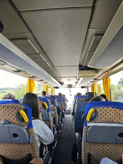 The image shows the interior of a bus traveling on a road, likely in the vicinity of Krivodol, Bulgaria, judging by the presence of rolling green hills and trees visible through the windows. The perspective is from the back of the bus, looking forward towards the front. The seats are upholstered with a brown and blue diamond pattern, with blue headrests. Several passengers are seated, with their backs to the camera. Yellow curtains are drawn over the windows. Above the seats, the ceiling of the bus features ventilation grilles and light fixtures. The aisle of the bus is visible, with luggage netting attached to the back of some seats. The overall impression is one of a typical daytime bus journey through a rural landscape.