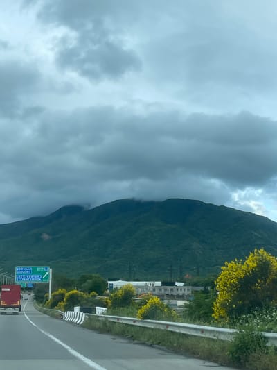 The image depicts a landscape scene in the city of Gorovani, Georgia. A mountain covered with dense green vegetation dominates the right side of the frame, its base partially obscured by thick, low-lying clouds. To the left, the sky is overcast with dark, heavy clouds, suggesting inclement weather.
A road runs along the base of the mountain, featuring a guardrail and a white line marking the lane. A road sign is visible on the side of the road, with the names of cities like Gudauri and Stepantsminda, written in both Georgian and English. A red vehicle is seen driving on the road.
In the middle ground, structures are visible including multi-story buildings. Yellow flowering trees are scattered along the roadside.