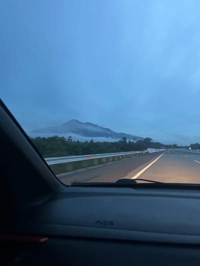 The image depicts a view from inside a vehicle, looking out at a highway in Seulimeum, Indonesia, at dusk or dawn. A mountain range shrouded in mist is visible in the distance, with lush green vegetation bordering the highway. The road is illuminated by the headlights of oncoming vehicles and streetlights. The sky is a deep, overcast blue. The interior of the car, including the dashboard and a portion of the windshield frame, is visible in the foreground, suggesting the perspective of a driver or passenger. There are no people or other animals visible in the scene. The overall mood is tranquil and slightly atmospheric due to the low light and misty mountains.