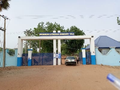 The image shows the entrance to the Bronnum Lutheran Seminary Yola, located in Rumde Waziri, Nigeria. It is a white, gated entrance with blue accents. The gate is open and there is a white car driving through it. A man is walking behind the car. The gate is adorned with a sign that reads "Bronnum Lutheran Seminary Yola." There are power lines running overhead and a building with a blue roof on the right side of the gate. The ground is dirt and there is vegetation in the background. The sky is clear and blue.