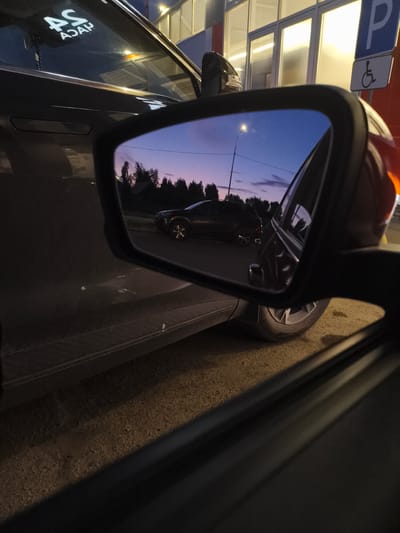 The image shows a view from inside a car looking out the driver's side window and into the side mirror. The time of day appears to be dusk, with a gradient of purple and blue in the sky.
In the side mirror, a black SUV is reflected. The SUV is parked on a paved surface, possibly a road or parking lot, with trees and a small building visible in the background. A street light is also reflected in the mirror, illuminating the scene.
Outside the car, the vehicle's dark gray paint is visible. On the rear window of the car, a sticker with white lettering that reads "AS ASAP" is applied. To the right of the car, a blue parking sign with a white wheelchair symbol is mounted on a pole, indicating a disabled parking spot. The building behind the sign has large windows with interior lights on.
The foreground shows the interior of the car, with the door frame and the edge of the car's interior panel visible. The ground beneath the car is rough and appears to be gravel or dirt. The image seems to be taken in a city or town in Russia, potentially near a building or parking area.