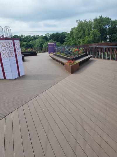 The image depicts an outdoor scene in the city of Kuršėnai, Lithuania, likely a public space or park. In the foreground, a wooden deck is visible, with a section of it in a lighter shade and another in a darker shade. To the left, a large white structure with maroon stripes and a decorative pattern is present, and to its left, large translucent numbers "30" are visible. Further into the scene, a planter box with red flowers sits on the deck. Behind the planter, there are benches and more wooden decking. A metal railing with mesh infill borders the elevated area. In the background, lush green trees form a dense treeline, and the sky is overcast with grey clouds. A purple informational banner with text and graphics stands near the planter box.