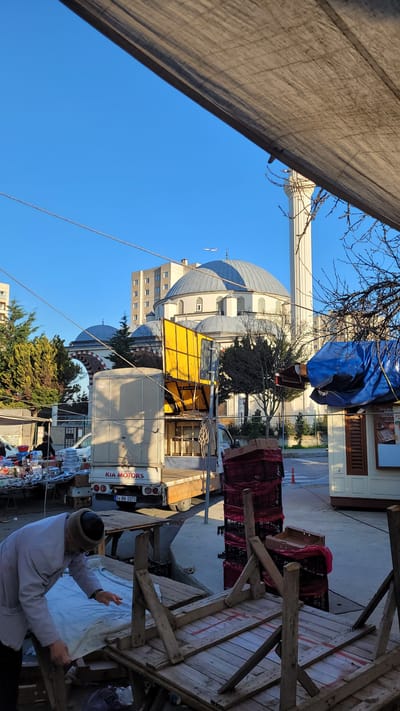 The image depicts a street scene in Beylikdüzü, Turkey, under a clear blue sky. A large mosque with multiple domes and a tall minaret is visible in the background. A white KIA Motors truck is parked on the street, partially obscured by trees. A wooden structure, possibly a market stall, occupies the foreground. A man wearing a light-colored jacket and a dark hat is standing near the stall, appearing to be arranging items on it. The overall scene suggests a marketplace or street vendor setting up for business.
