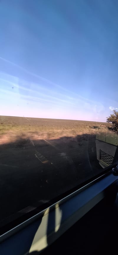 The image captures a landscape view, likely from a moving vehicle, with a clear blue sky dominating the upper portion. A vast, flat expanse of earth, appearing as cultivated fields or barren land, stretches across the middle ground, exhibiting a motion blur that suggests forward movement. Towards the right edge of the frame, a cluster of trees is visible, also blurred by motion. In the foreground, a dark, reflective surface, likely a window, shows some smudges and reflections. Part of what appears to be the interior of a vehicle is also visible along the bottom edge. The scene does not provide any specific landmarks or details that would allow for definitive identification of the location within the city of Morava, Bulgaria.