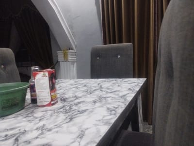 In a dimly lit room in Iyafin, Nigeria, a marble-patterned table dominates the foreground. On the table sits a green basin, a bottle of dark liquid, and a red and white box with text and an image of a woman's face. A small, round, purple object rests near the box. Behind the table, two dark upholstered chairs are visible. The room is furnished with draped brown curtains and white architectural trim. The overall impression is one of a domestic or commercial setting, possibly a shop or a home, with items suggesting everyday use.