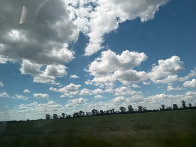 The image shows a wide, flat landscape under a bright blue sky dotted with fluffy white clouds. In the foreground, a dark green field stretches horizontally across the frame, appearing slightly blurred, suggesting movement. A line of trees, mostly silhouetted against the sky, forms a dark border along the horizon, separating the field from the sky. The clouds are varied, with some appearing dense and cumulus-like, while others are thinner and more wispy. The overall impression is of a rural setting on a clear, possibly breezy day. The image does not contain any people, buildings, or urban elements.