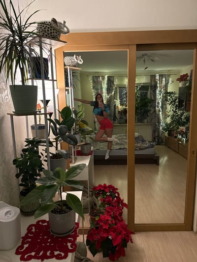 A mirror shows a room interior with a bed against a wall adorned with fairy lights and patterned curtains. A woman in a sports bra and shorts is posing in a handstand in front of the mirror. In the foreground, a shelving unit holds an array of potted plants of different sizes and species, with a red poinsettia on the right. Decorative items, including a spotted elephant figurine, also sit on the shelves.