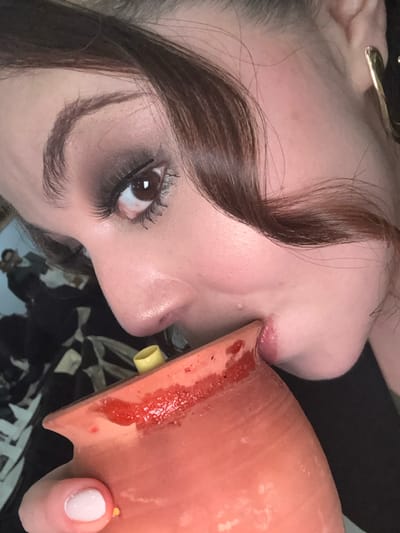 A fair-skinned woman drinks from a reddish-brown pottery cup. The cup rim touches her lips. A small, yellow straw-like object is visible on the cup's surface near her nose. She has dark brown hair with a prominent curl falling across her forehead. Her eye is visible, lined with dark eye shadow and mascara. She has a visible ear with a gold earring. Her fingernails are painted white.