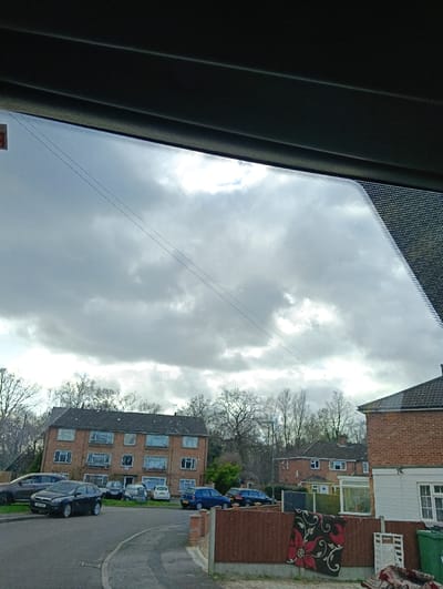 This image presents a residential street view in Surrey Heath, United Kingdom, captured from inside a vehicle through a windshield displaying small water droplets or smudges. The sky is predominantly overcast with areas of brighter cloud cover.

In the immediate foreground, the dark grey interior structure of a vehicle partially obstructs the top of the frame. A paved road or driveway curves left from the bottom center. A low brown fence with brick posts runs along the right side of the foreground. Draped over this fence is a dark rug featuring a prominent red floral or swirling pattern. Behind the fence, on the far right, are two green wheeled bins and a white lattice panel.

The midground features a three-story, multi-windowed brick apartment building on the left, with a dark roof. Several vehicles are parked in front and around this building, including a dark grey hatchback on the road in the left foreground, a dark blue hatchback, and various lighter-colored cars. To the right, a red-brick house with a white ground-floor extension and a tiled roof is visible. Numerous bare deciduous trees, indicating a dormant season, are scattered throughout the background and behind the structures, alongside some evergreen foliage. Two thin, dark power lines are visible against the sky, extending across the scene. No individuals are present.