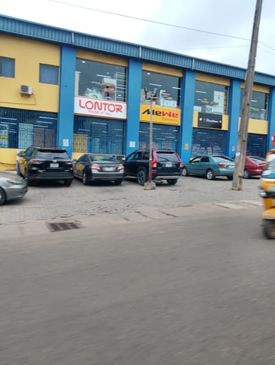 The image shows a street scene in front of a commercial building with a bright yellow facade and blue columns. Several cars are parked in front of the building. A sign on the building reads "LONTOR POWER AT WILL". Another sign displays "MeWe Electronics". A third sign advertises "BinaTone". To the right of the building, a yellow auto-rickshaw is visible. The ground is paved with interlocking bricks. The sky is overcast. The scene appears to be a commercial area in Isheri, Nigeria.